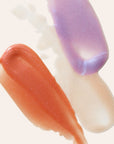Close up of peach lavender and iridescent lip gloss swatches on a pale background