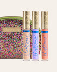 Three LipSense gloss tubes with ornate gold caps in front of a multicolored glitter clutch